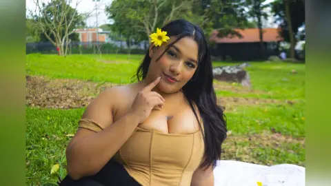 Smiling woman in a tan corset top sitting on grass with a yellow flower in her hair in a sunny garden.