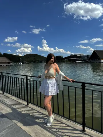 Young woman in a white crochet bikini top and skirt posing by a lake under a sunny blue sky.