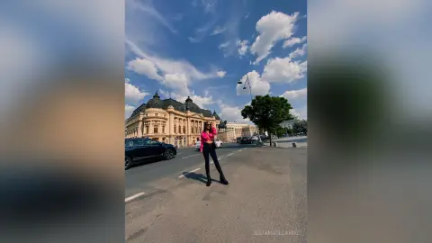 Stylish woman in a pink top and black jeans standing on a city street under a bright blue sky.