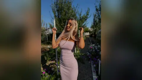 Anita stands outdoors in a tight, strapless light purple dress with her blonde hair down.