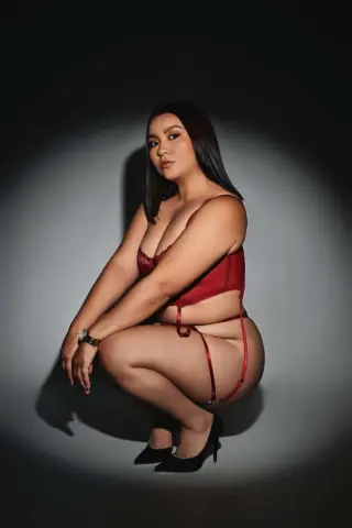 Curvy woman in elegant red lace lingerie posing against a dark studio background with dramatic lighting.