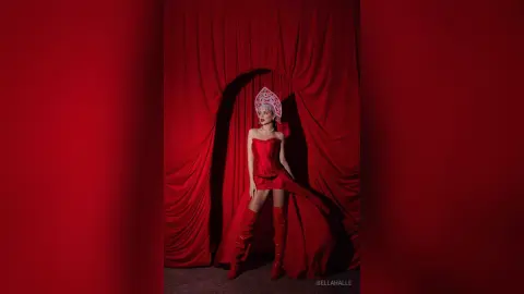 Elegant woman in a red couture gown and tall boots posing against dramatic red velvet curtains.