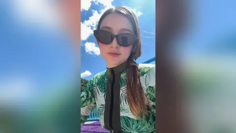 Young woman in green tropical print shirt and sunglasses smiling under a bright blue sky.