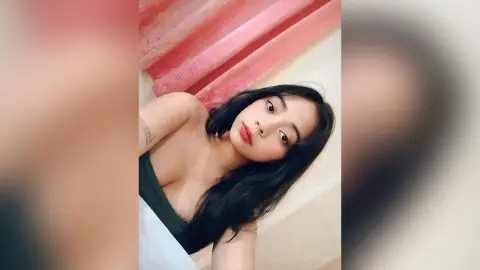 Young woman in a dark green top posing with a soft, natural look against a pink curtain background.