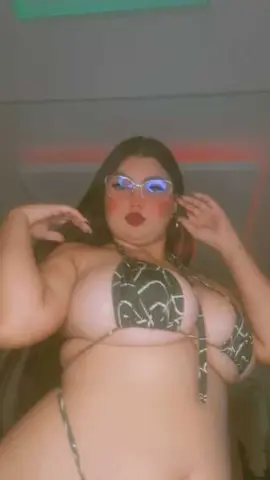 Curvy woman in a patterned bikini posing confidently in a dimly lit indoor setting.