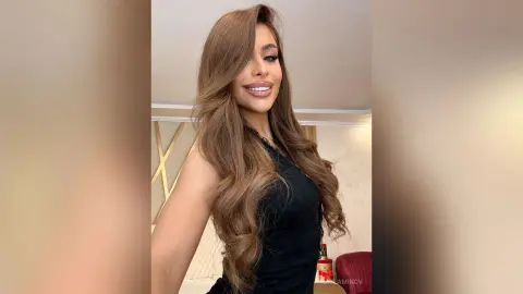 Smiling woman with long wavy brown hair wearing a black sleeveless top in a bright indoor setting.