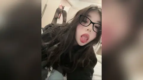 EltonMia wears black glasses and sticks her tongue out while posing in a dark sweater.