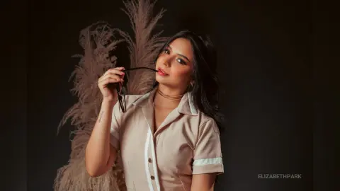 Elegant woman in a beige button-down shirt posing with sunglasses against a dreamy pampas grass background.