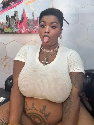 Tattooed woman in a white ribbed top sitting playfully in front of a city skyline painting.