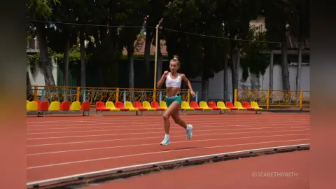 Athletic woman in white sports bra and green shorts running on a red outdoor track under bright daylight.