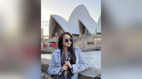Stylish woman in a light blue jacket and sunglasses smiling in front of the Sydney Opera House at sunset.