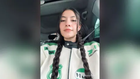 Young woman with long dark braids wearing a white and green sporty jacket sitting in a car.