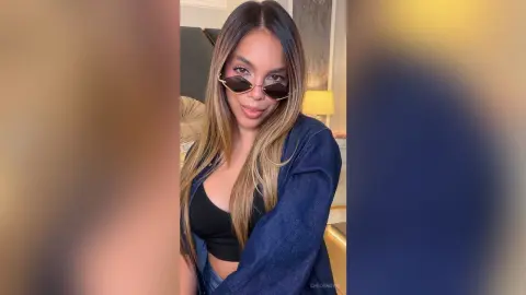Smiling woman in a black crop top and blue denim jacket wearing stylish sunglasses in a cozy indoor setting.