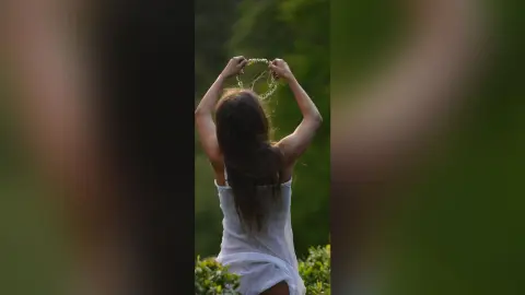 Young woman in a white summer dress holding a flower crown in a lush green meadow at sunset.