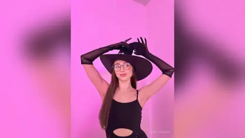 Young woman in black tank top and wide-brimmed hat posing against a vibrant pink background.
