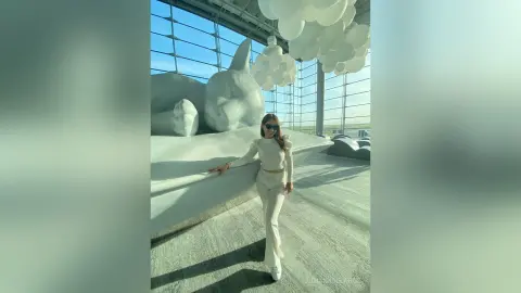 Stylish woman in white trousers and long-sleeve top standing in a bright, modern minimalist art gallery.