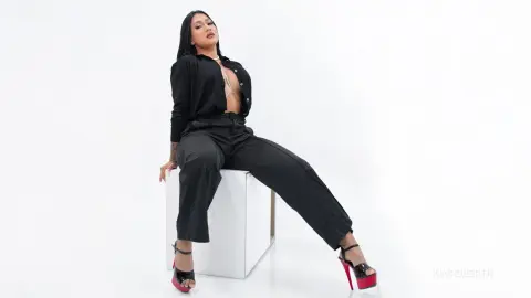 KimRoberth sits on a white cube wearing a black button-down shirt and black trousers with red high heels.
