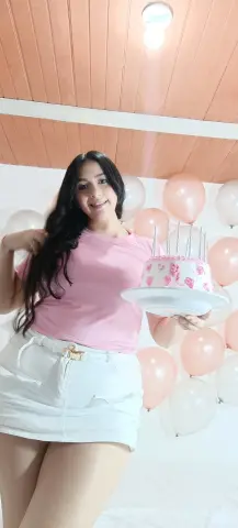 Smiling young woman in pink t-shirt and white skirt holding a pink birthday cake with balloons in background.
