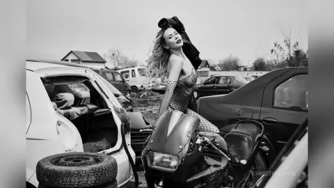 ChristineDavis poses outdoors next to a motorcycle and wrecked cars, wearing a black fishnet bodysuit with long sleeves and long blonde hair.