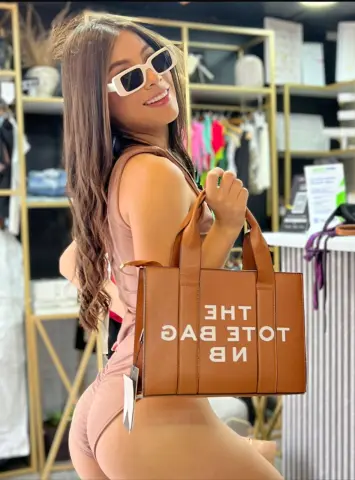 Smiling woman in a tan bodysuit holding a brown THE TOTE BAG in a bright clothing boutique.