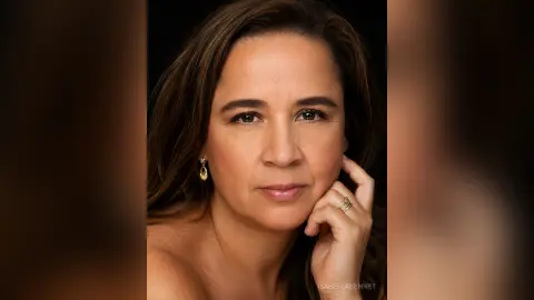 IsabellaBennet poses for a close-up headshot, resting her chin on her hand with long brown hair and gold earrings against a dark background.