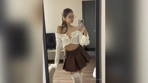 EmorySwift poses in a mirror wearing a white off-the-shoulder lace-up crop top and a short brown pleated skirt with white thigh-high socks.