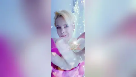 AvaLindt with blonde hair poses in a pink top, blowing a kiss toward the camera against a soft, glowing background.