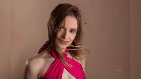 Gia poses with her long brown hair swept to the side, wearing a bright pink halter-style top against a plain tan background.