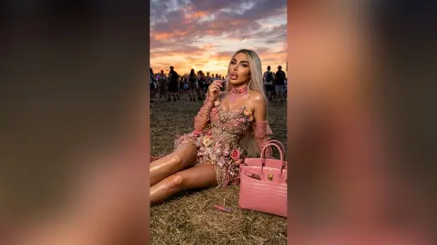 Glamorous woman in a pink floral mini dress sitting in a field during a dreamy sunset outdoor festival.