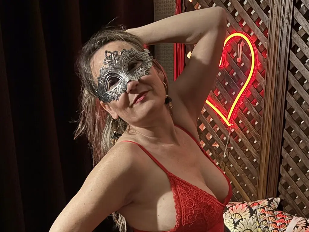 Woman in a red lace bodysuit wearing a silver masquerade mask in a moody, neon-lit lounge setting.
