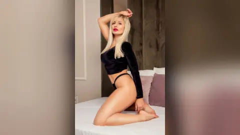Gia poses naked on a white bed, wearing a black velvet long-sleeve crop top and black thong, running her hand through her blonde hair.