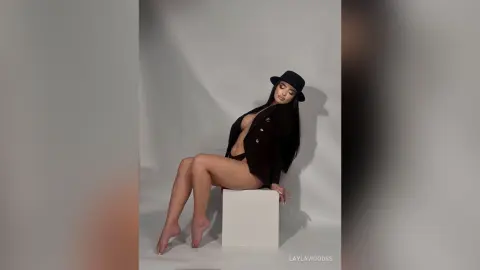 LaylaWoodss sits on a white block wearing a black hat and a black open jacket with black underwear, showing her long dark hair and bare legs.