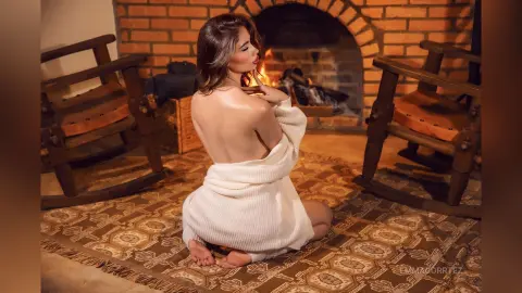 Woman in white knit sweater sitting on a patterned rug by a cozy glowing fireplace in a rustic room.