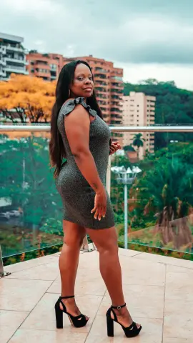 Confident woman in a grey sleeveless mini dress posing on a balcony overlooking a lush tropical cityscape.