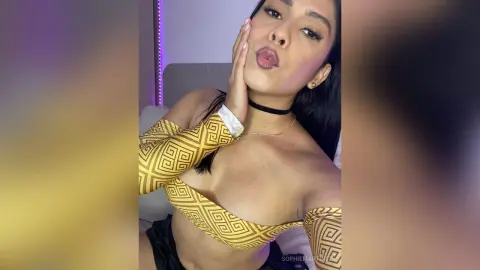 Young woman in a yellow patterned crop top posing confidently in a bedroom setting.