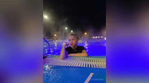 Young man in a black shirt relaxing in a glowing blue night pool under misty lights.