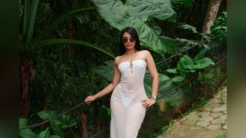 Woman in a white sheer polka dot dress and sunglasses posing in a lush tropical garden setting.