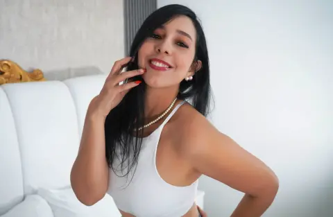 KattyRivera — Smiling young woman in a white tank top and pearl necklace relaxing in a bright bedroom.