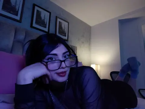 Young woman with glasses in a black long-sleeve top relaxing on a bed in a moody, dimly lit bedroom.
