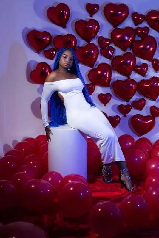 Hanna_Hall poses with her long blue hair, wearing a white off-the-shoulder top while posing against a red background.