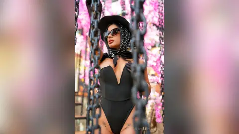 Stylish woman in a black bodysuit, sunglasses, and headscarf posing amidst pink floral decorations.