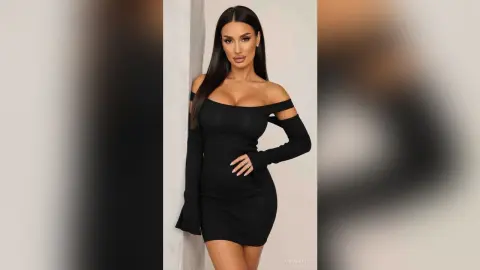 AnnaKarev poses in a tight black off-the-shoulder mini dress with long sleeves against a plain wall.