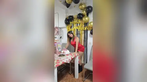 Happy woman in a red dress celebrating her birthday with a cake and gold and black balloons at a party table.
