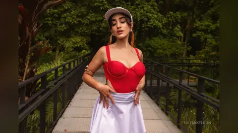 Young woman in a red bodysuit and white skirt posing on a garden walkway surrounded by lush green foliage.