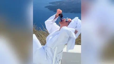 MissyJolie wears a white towel wrap and sunglasses while eating dark grapes overlooking a blue ocean coastline.