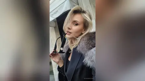 Blonde woman in a black blazer and grey fur coat posing elegantly in an urban city setting.