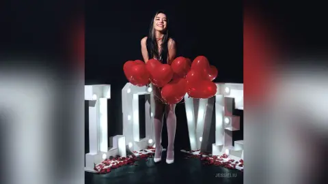 JessieLiu smiles while standing behind a row of red heart balloons, wearing a black lace bodysuit against a black background.