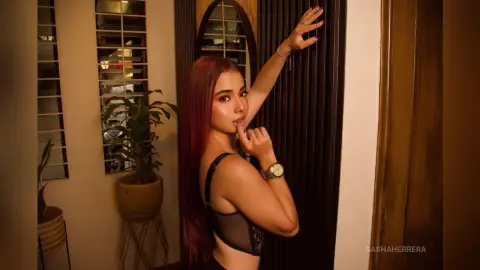 Stylish woman with long red hair in a black bra posing confidently in a warm, moody indoor setting.