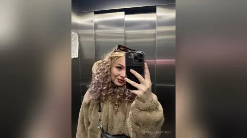 Young woman with curly blonde hair in a cozy brown fuzzy jacket taking a selfie in a modern elevator.