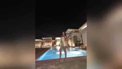 NicoleBella stands poolside at night wearing a short, tight gold dress and green heels.